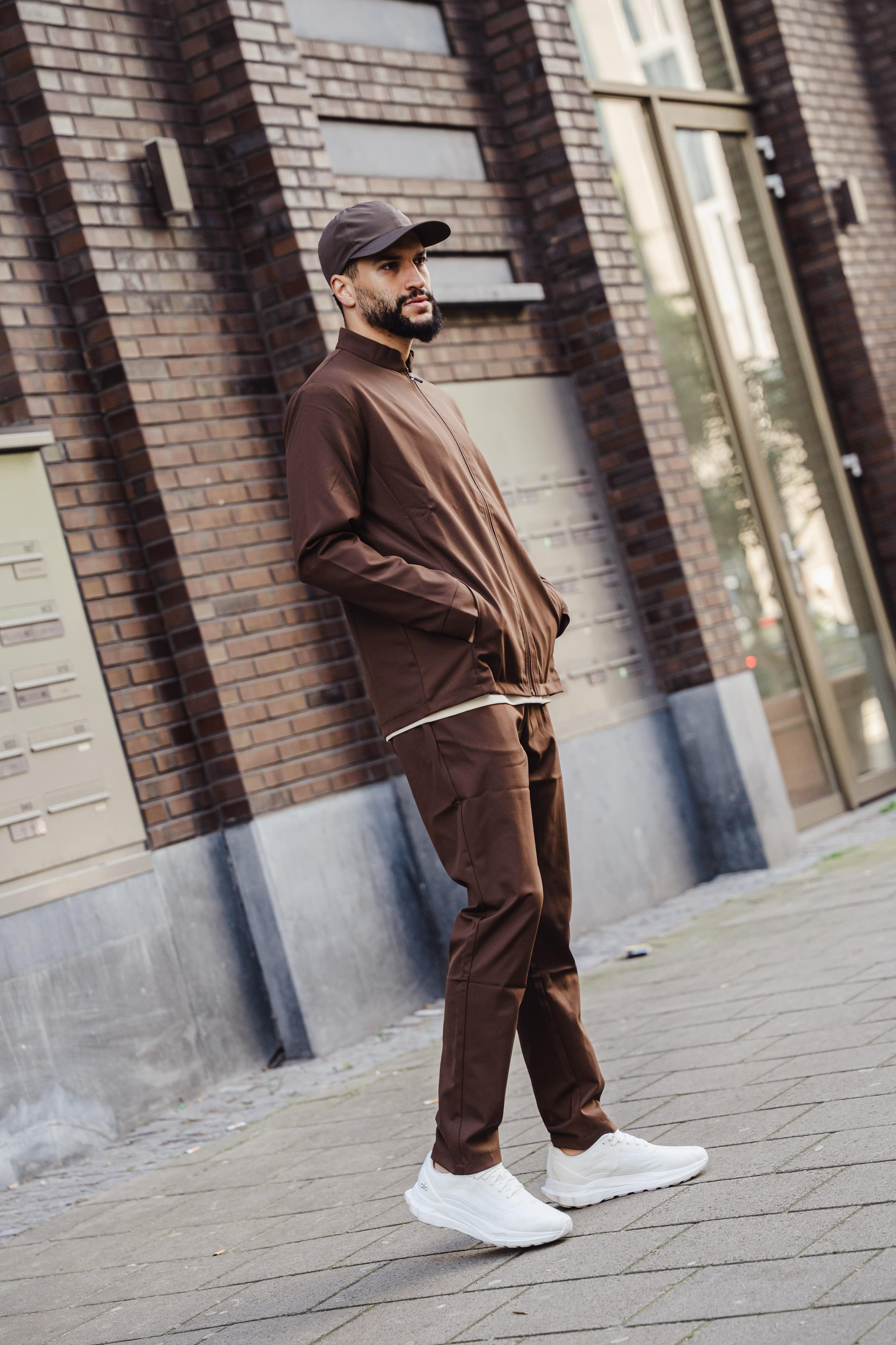 Man in een bruin tracksuit en pet die op straat tegen een bakstenen gebouw leunt, met witte sneakers aan.