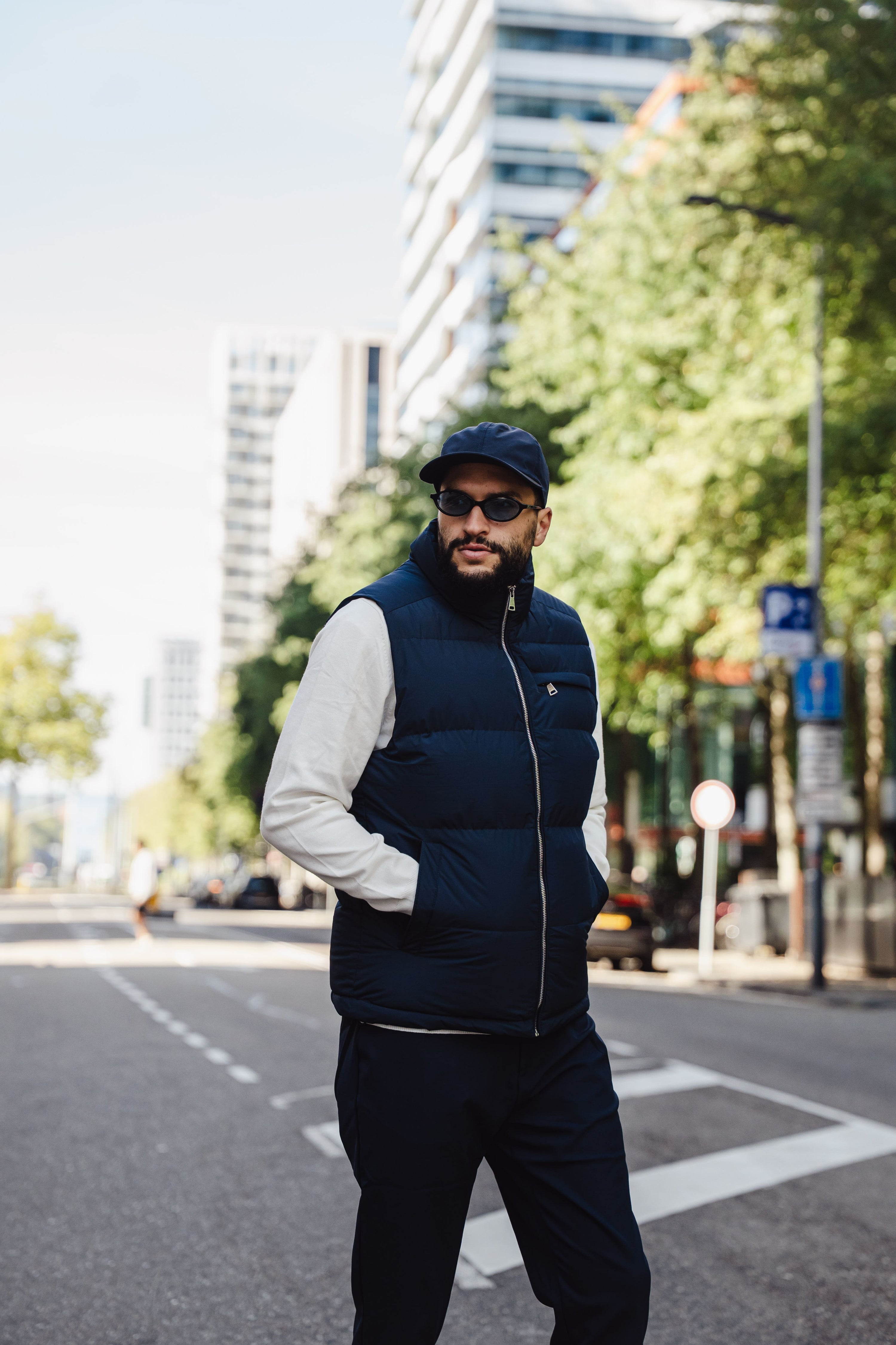 Man die over een stadsstraat loopt, gekleed in een donkerblauwe gewatteerde bodywarmer, lichte trui, donkere broek, zonnebril en pet, met hoge gebouwen en bomen op de achtergrond.